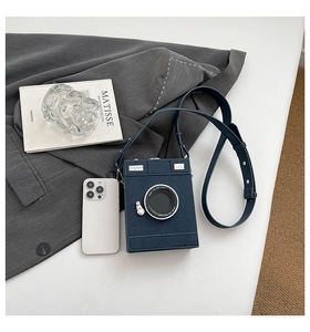 Detail of camera-shaped crossbody bag with lens design next to a smartphone for size reference