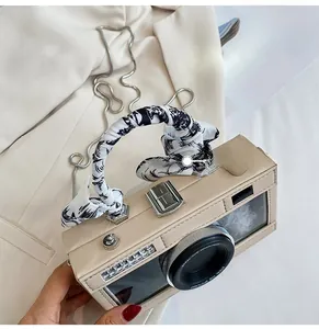 Detail of camera-shaped bag with scarf-wrapped handle and metal chain on neutral background