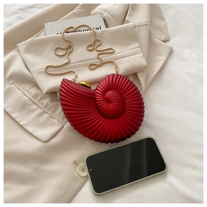 Close-up of a burgundy conch-shaped acrylic box bag with a gold chain strap on fabric