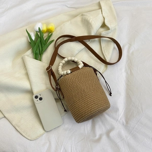 Woven straw bucket bag with white wooden bead handle and brown leather shoulder strap