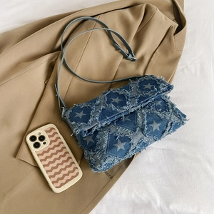 Blue denim crossbody bag with white star prints and trendy frayed edges on khaki backdrop