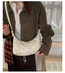 Close-up of a beige hobo bag with silver rivets worn over the shoulder in a cozy indoor setting