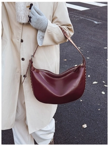Detail shot of a dark red leather crescent bag with silver rivets worn over a beige trench coat