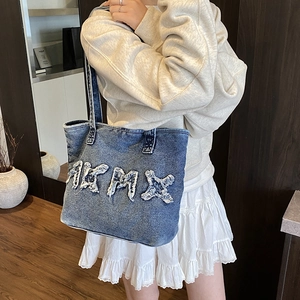 Blue denim tote bag with frayed AKMX letter design shown on model in a minimalist room