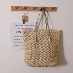 Minimalist beige woven straw tote bag with slim leather straps hanging on a wooden hook