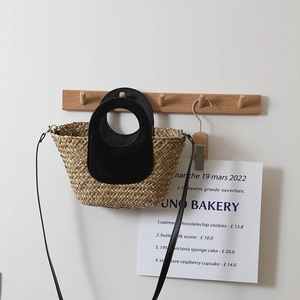 Handwoven straw crossbody bag with black leather trim hanging on a wooden hook