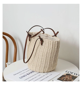 Close-up of a handwoven straw bucket bag with slim brown leather handles on a white table