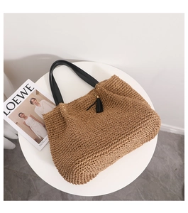 Detail of woven straw tote bag with black leather straps and tassel on a white table
