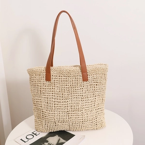 Large beige woven straw tote bag with brown leather handles on a white minimalist table
