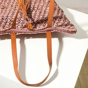 Pink woven straw tote bag with brown leather handles and decorative tassel for beach vacations