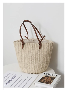 Detail of hand-woven straw bucket bag with brown leather handles on a white table