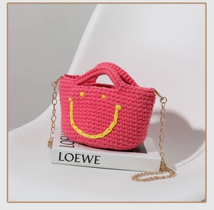 Sac seau en maille rose avec smiley jaune et chaîne dorée posé sur un livre blanc
