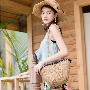 Straw tote bag with black pom-poms and long straps in a natural outdoor setting