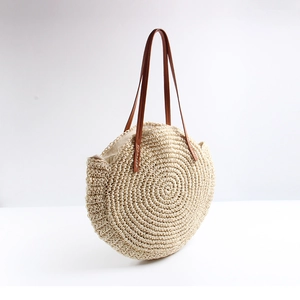 Close-up of beige round straw tote bag with brown leather shoulder straps on white background