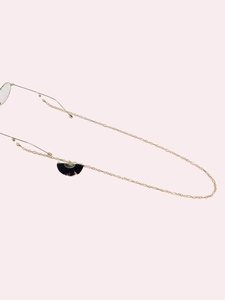 Detail of gold metal eyewear chain featuring a black fan tassel on a soft pink background