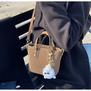 Brown felt bucket bag with a cute white plush monster charm shown on a park bench