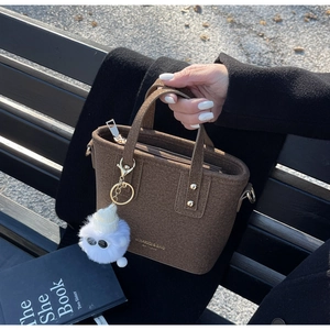 Detailed texture of brown felt bucket bag with top handle and a white fuzzy pompom charm
