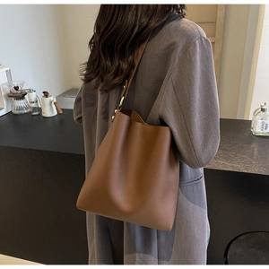 Brown litchi grain leather bucket bag shown on shoulder in a minimalist indoor setting