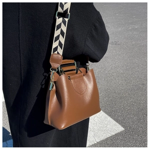 Detail of a brown bucket bag featuring a wide geometric woven strap and gold metal handle