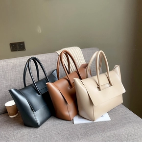 Three minimalist matte leather tote bags in black, caramel, and beige on a grey sofa