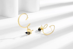 Détail de boucles d'oreilles abeille dorées avec courbe en C sur fond géométrique blanc