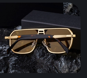 Macro view of gold metal square sunglasses with amber temples on a black rock surface