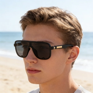 Close-up of black shield sunglasses with gold metal accents on a male model at the beach