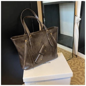 Detail view of large brown tote bag featuring decorative zippers and long tassels on white box
