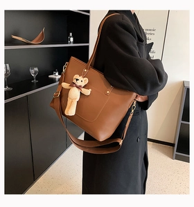 Detail of a brown leather tote bag featuring a cute knit bear charm and wide strap