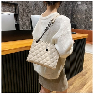 Beige quilted shoulder bag with chain strap shown on model in a modern interior setting