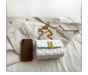 Close-up of white quilted crossbody bag with gold bunny ears lock next to a phone