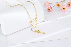 Close-up of golden necklace featuring a hollow heart and key pendant on a white stand