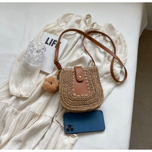 Détail du sac bandoulière en paille avec rabat en cuir brun à côté d'un smartphone pour la taille