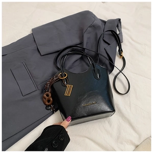 Detalle de bolso trapezoidal negro con logo dorado, correa trenzada y dije abalorio