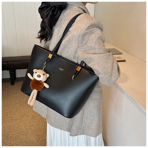 Large black leather tote bag with plush bear charm worn by model in a stylish indoor setting