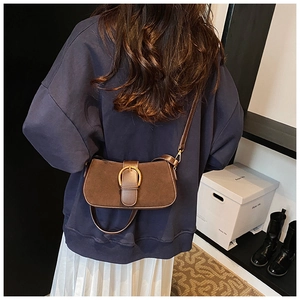 Model wearing dark brown crossbody bag with gold buckle in minimalist indoor setting