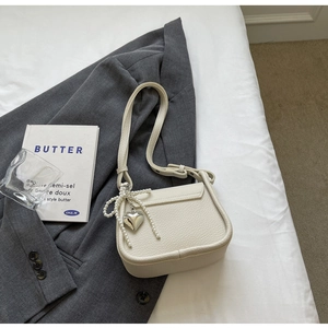 Close-up of beige lychee leather saddle bag with heart charm and pearl bow on white bedding