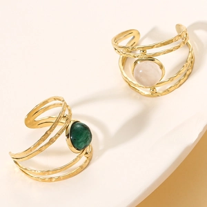 Detail of gold-plated multi-layer wavy rings with green and pink gemstones on cream stands