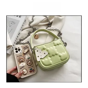 Grüne Mini-Handtasche mit Bärenprägung und Kettenriemen im Größenvergleich zum Handy