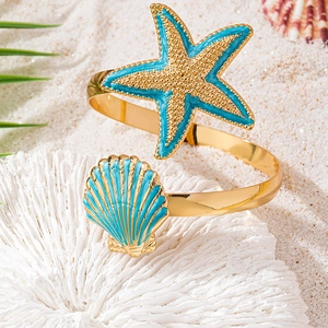 Golden open starfish bangle with blue enamel seashell charms on white beach sand