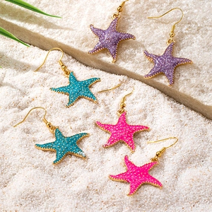Three pairs of colorful starfish earrings in purple, green, and pink on beach sand