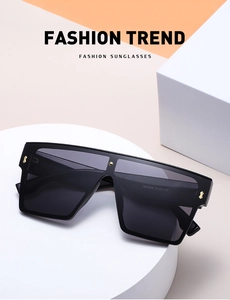 Detail of oversized black flat top sunglasses with gold rivet accents on white podium