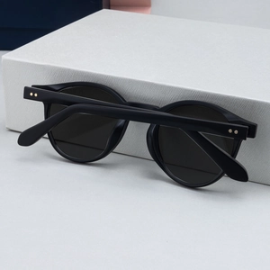 Detail view of round matte black sunglasses with golden rivet accents on white box