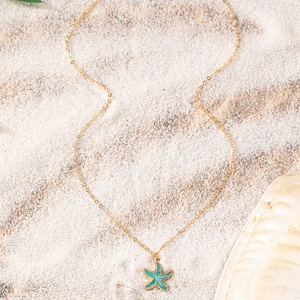 Detailed view of starfish pendant with blue enamel and gold chain on white beach sand