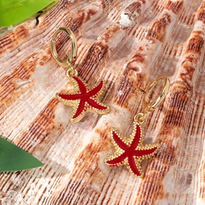 Golden starfish drop earrings with red enamel and huggie hoops on a seashell backdrop