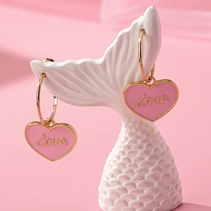Orecchini a cuore rosa con incisione Love appesi a un supporto in ceramica bianca