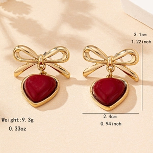 Elegant gold bow earrings with deep red heart pendants shown on a beige sand background
