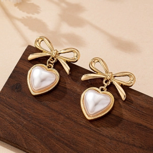Elegant gold bow earrings with heart-shaped white faux pearls on a vintage wooden surface