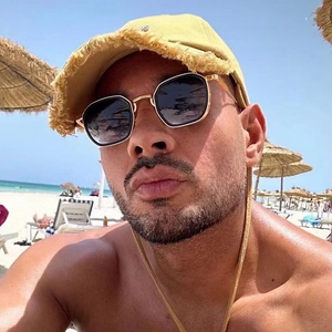 Men's gold hexagonal sunglasses with dark lenses shown in a beach resort setting