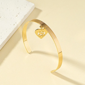Gold open bangle featuring a heart pendant with a cutout S letter in minimalist style